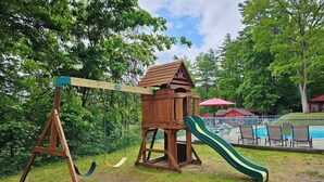 A wooden swing set with a slide, surrounded by grass and trees.