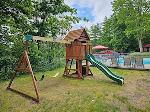 A wooden swing set with a slide, surrounded by grass and trees.