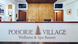 Un comptoir d’accueil à l’hôtel Pohorje Village Wellness & Spa, avec un mur en panneaux de bois, deux téléviseurs et un logo.