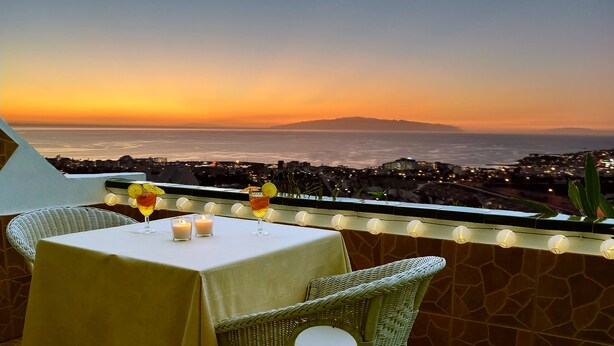 A table with drinks and candles on a balcony overlooking a cityscape at sunset.