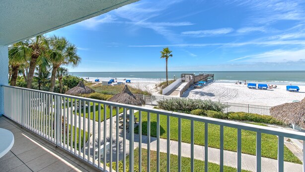 A beach view from a balcony with palm trees, a thatched-roof structure, and a sandy beach with blue lounge chairs.