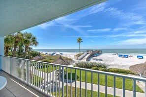 Uitzicht op een strand met palmbomen, een rieten gebouw en een zandstrand met blauwe ligbedden vanaf een balkon.