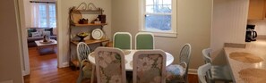 A dining area with a round table and floral chairs, a bookshelf with decorative items, and a view of a living room with a sofa and a coffee machine.