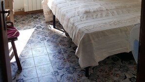 A bedroom with a bed, a window with a view of a building and trees, a radiator, a table with a lamp, and patterned floor tiles. - 2-storey vacation home with 2 large terraces and sensational views (Mazzano Romano)