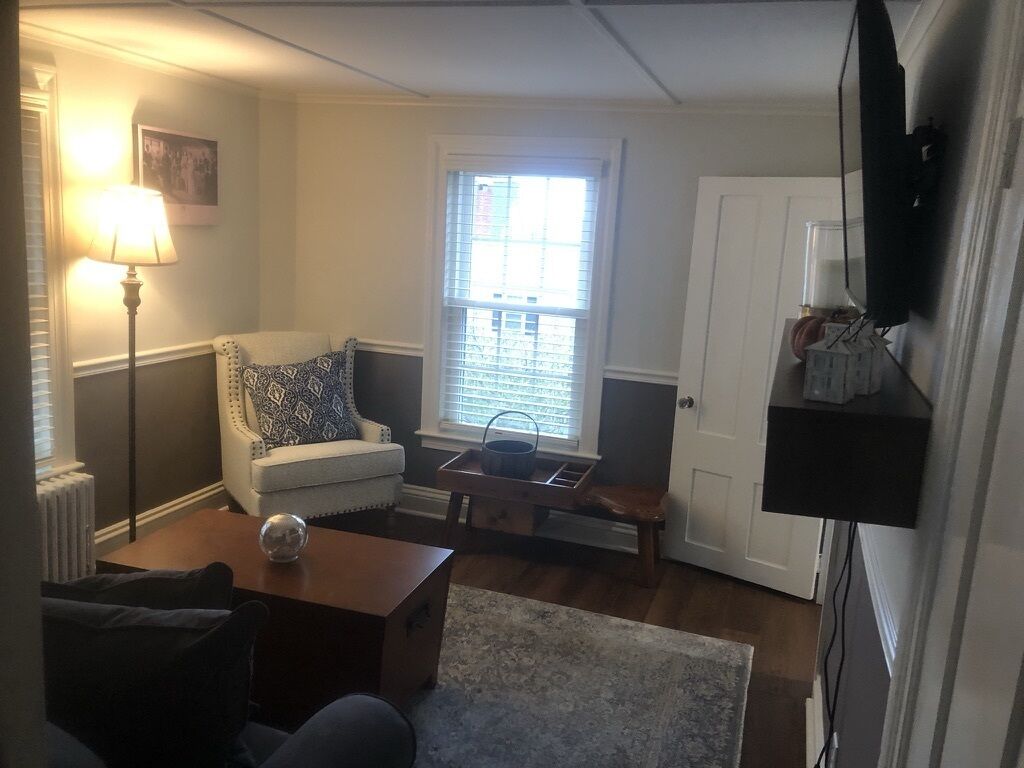 Een woonkamer met een bank, een salontafel, een lamp, een raam met zonwering en een televisie die aan de muur is bevestigd.
