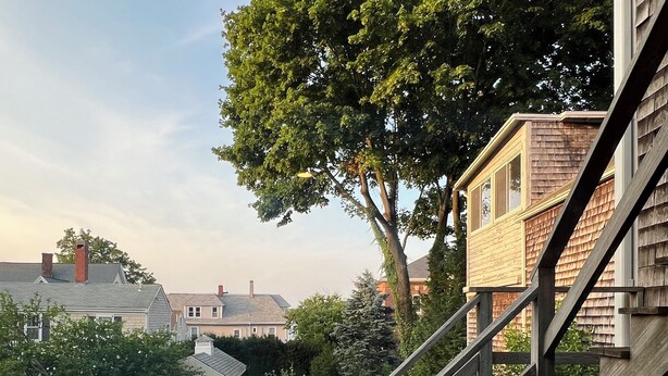 Una propiedad residencial con una escalera de madera que lleva a una terraza, rodeada por un patio cercado con un árbol grande y una casa pequeña al fondo.