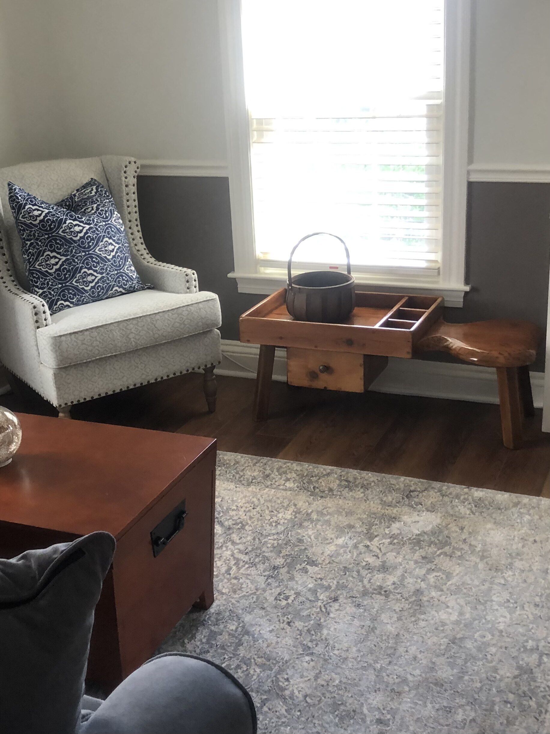 A living room with a white armchair, a wooden side table with a pot, a grey patterned rug, and a window with blinds.