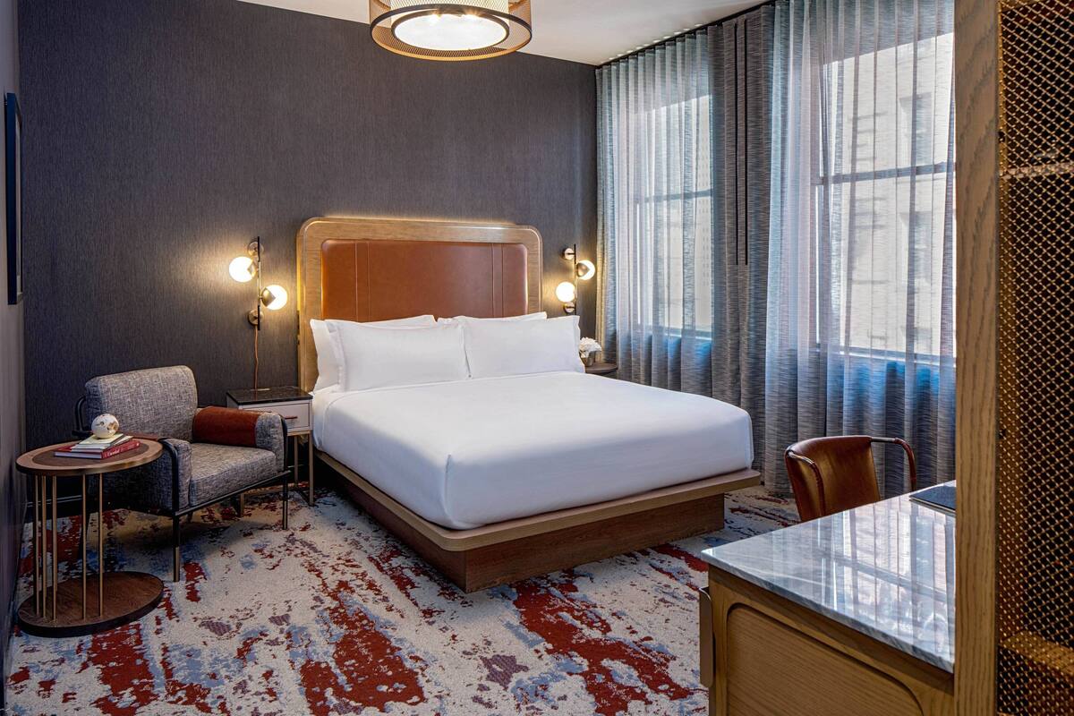A hotel room with a large bed, a small armchair, a desk, and a view of the city through large windows.