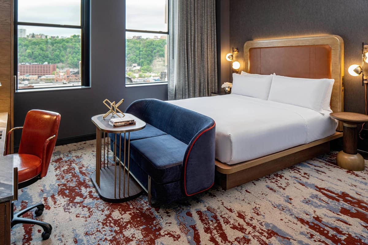 A hotel room with a large bed, a small table with a chair, a red chair, and a view of the city through large windows.
