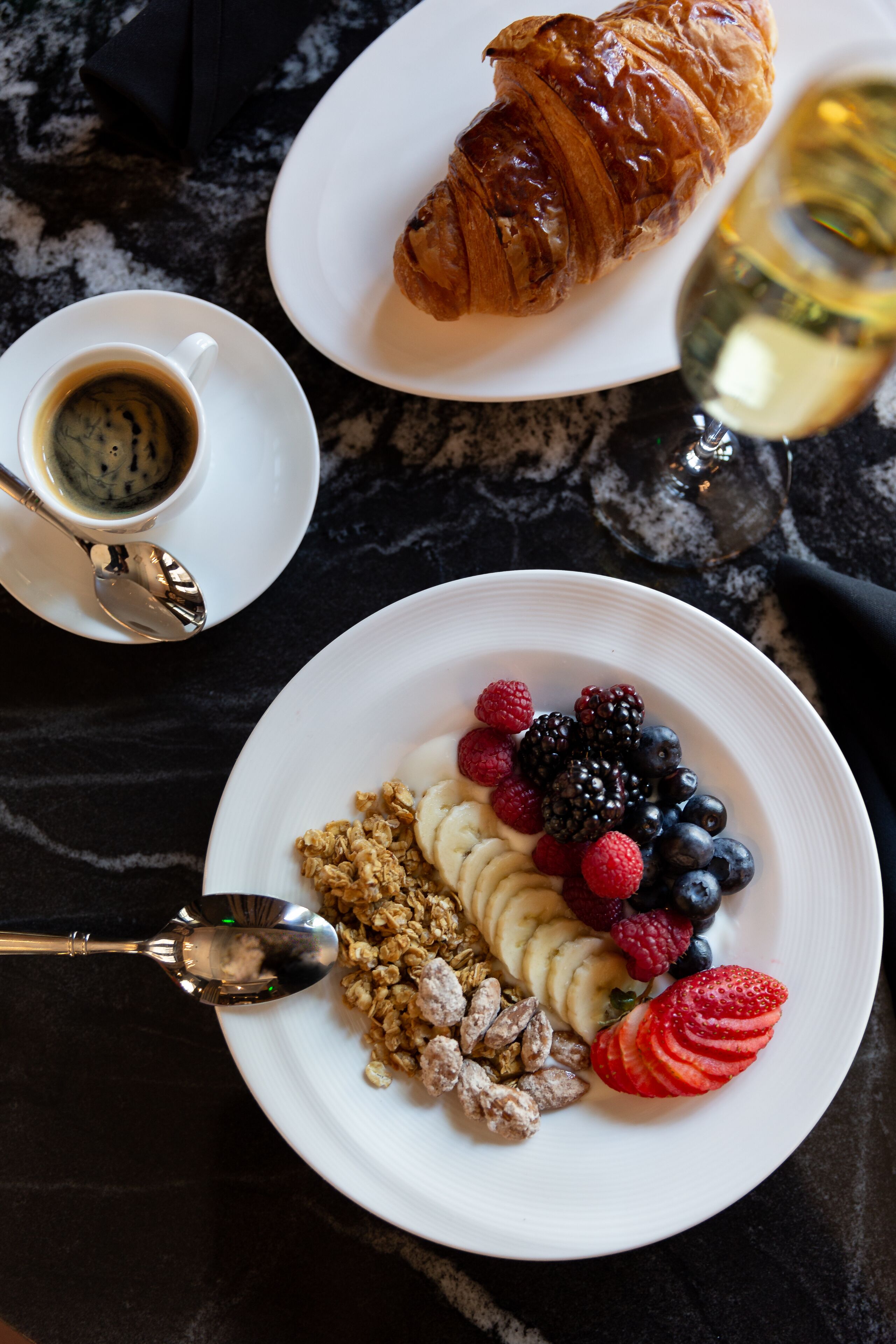 A breakfast setting with a croissant, a glass of white wine, and a plate of yogurt with fruits and granola.