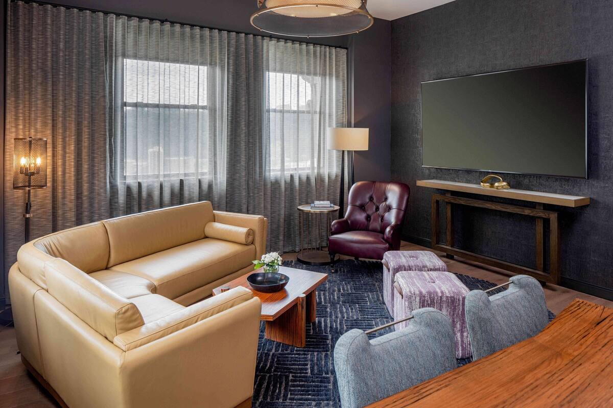 A modern living room with a beige sofa, a leather armchair, a wooden coffee table, and a TV mounted on the wall.