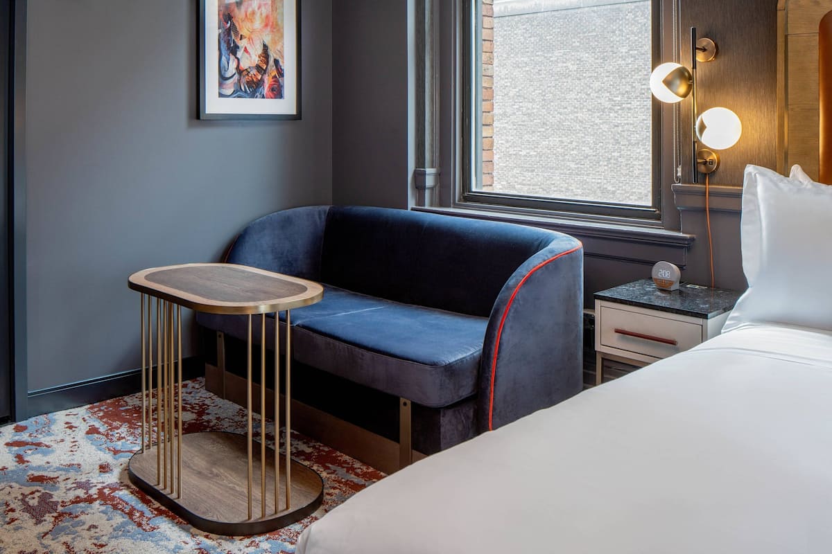 Ein modernes Hotelzimmer mit einem blauen Sofa, einem kleinen Holztisch, einem Bett, einem Nachttisch, einer Lampe und einem Fenster mit Blick auf die Stadt.
