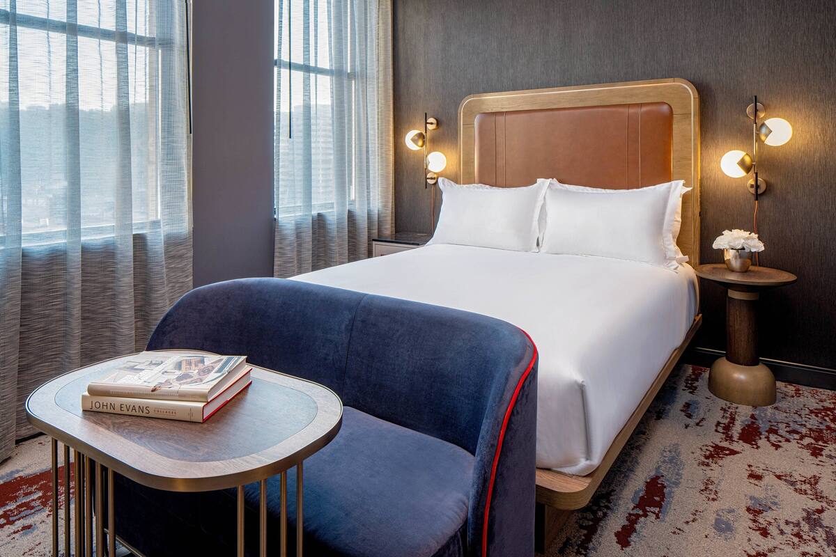A hotel room with a large bed, a blue armchair, a small round table with a book, and a nightstand with a lamp.