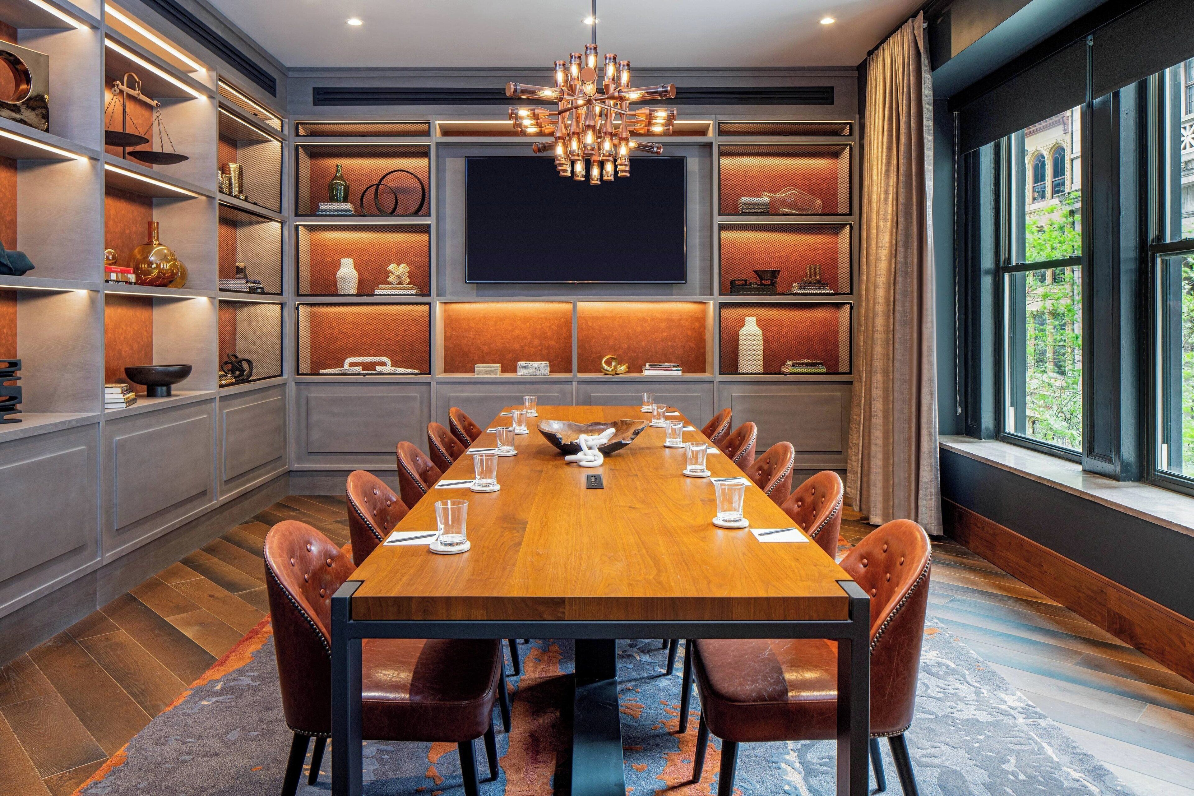 A conference room with a long wooden table, leather chairs, a chandelier, and shelves with various items.