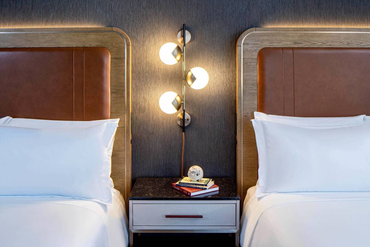 A hotel room with two beds, a nightstand with a lamp, and a small table with books and a decorative item.