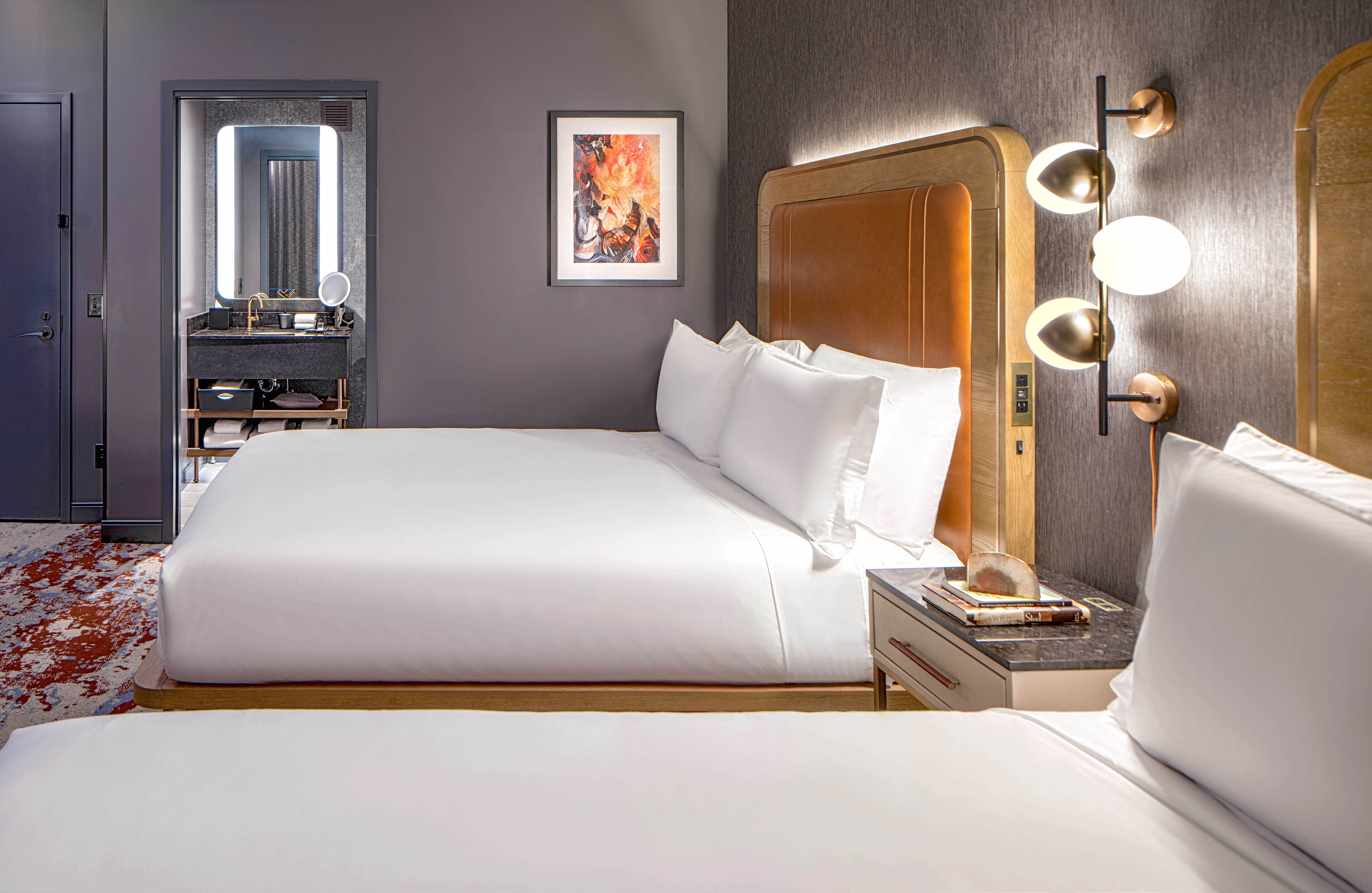 A modern hotel room with a large bed, a vanity with a mirror, and a wall-mounted light fixture.