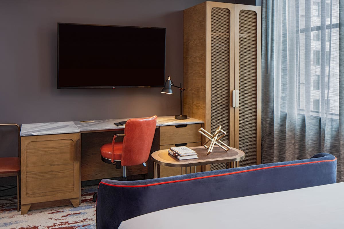 Ein Hotelzimmer mit einem Bett, einem Schreibtisch, einem Stuhl, einem Fernseher und einem Fenster mit Vorhängen.