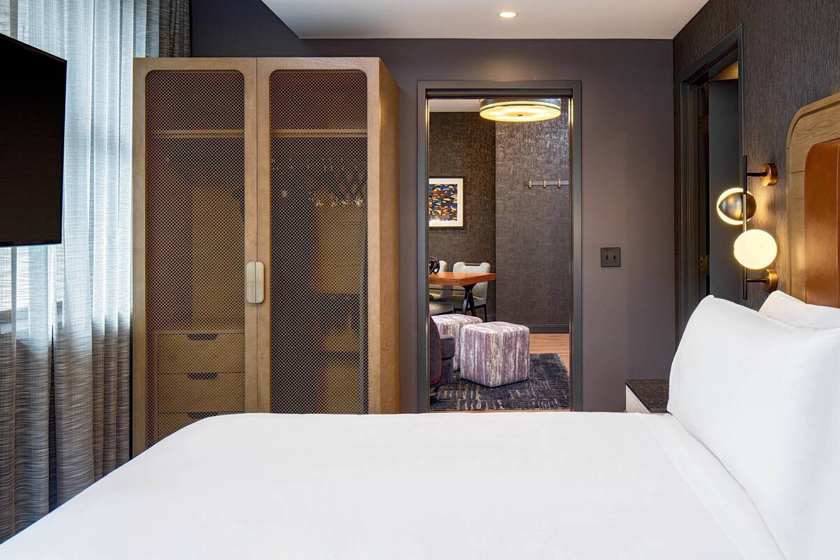 A hotel room with a large bed, a wooden wardrobe, a bathroom with a sink and mirror, and a television on the left.