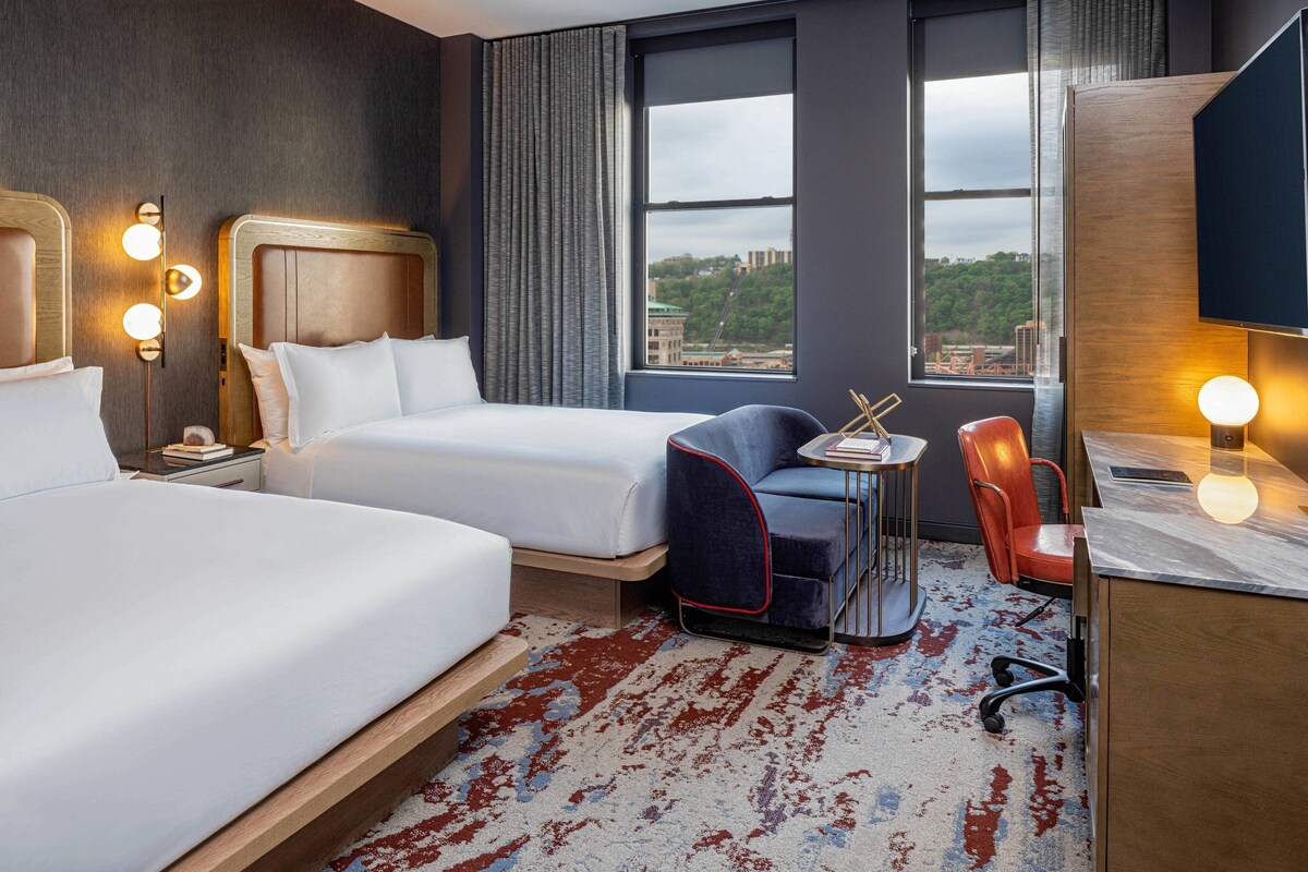 A hotel room with two beds, a desk, a chair, a small table, and a view of the city.