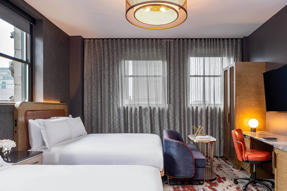 A hotel room with two beds, a desk, a chair, a lamp, and a large window with curtains.