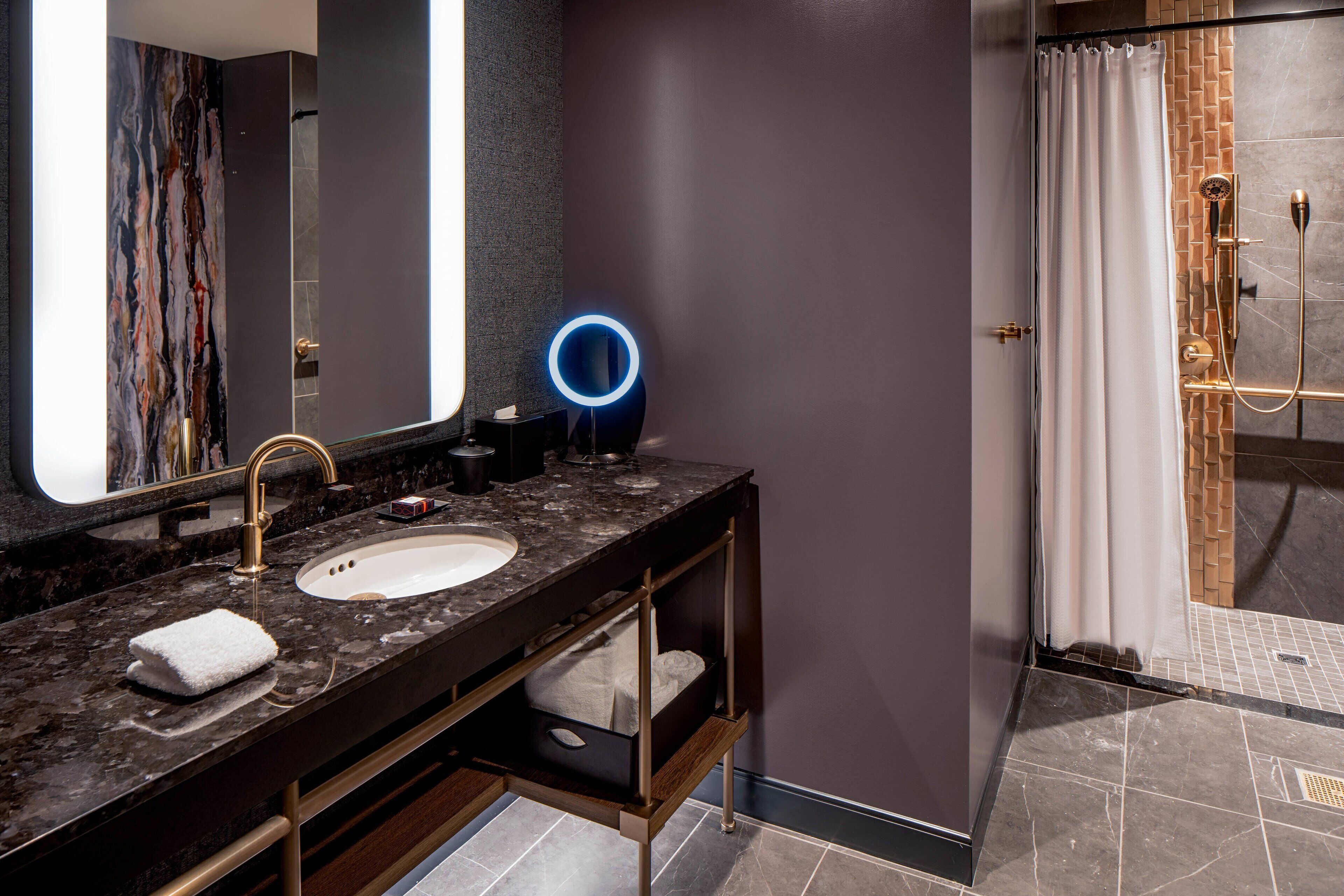 A modern bathroom with a marble sink, a large mirror, a blue illuminated ring light, and a shower area with a glass door.