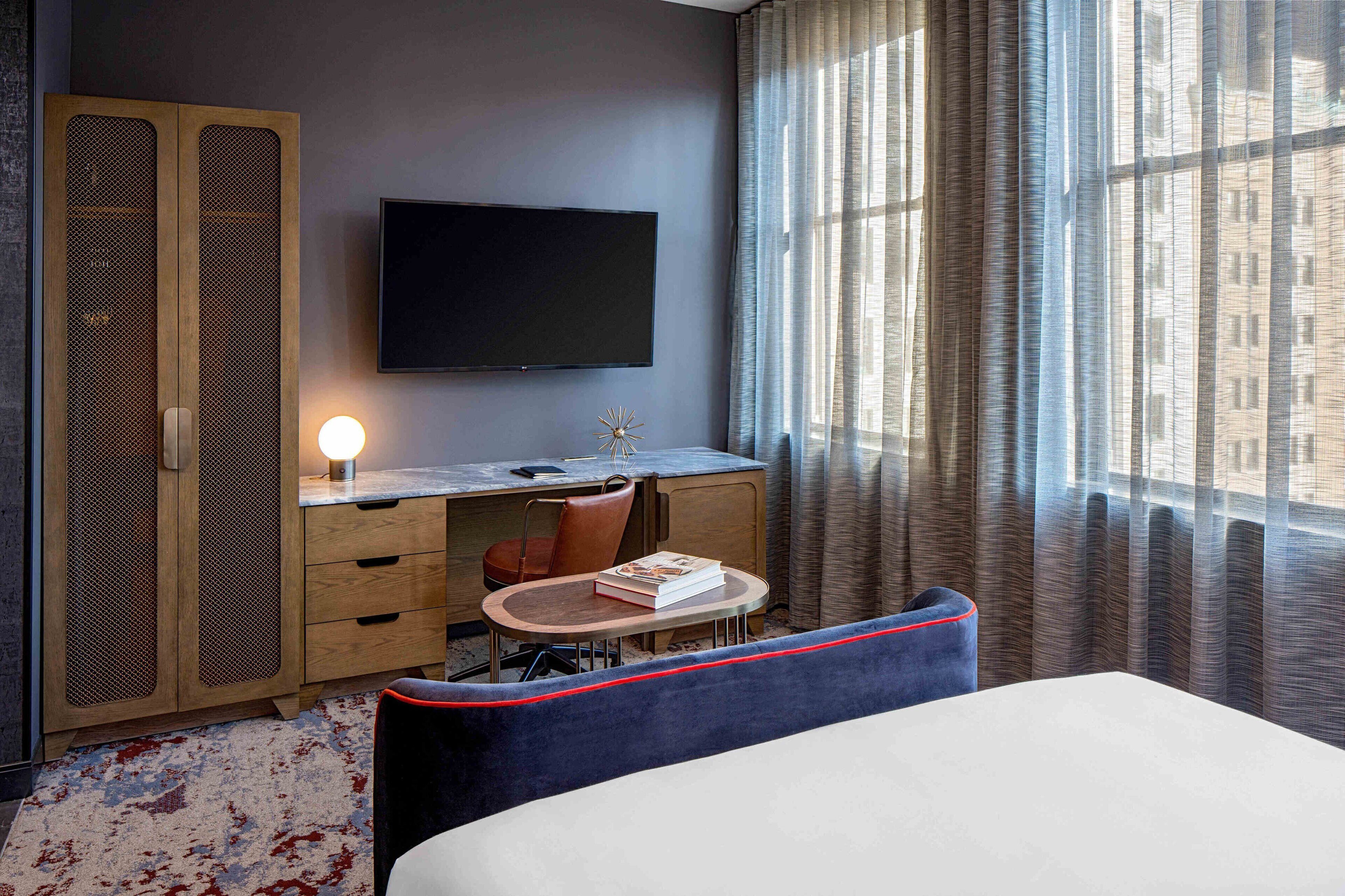 A hotel room with a bed, a desk with a chair, a television, and a window with curtains.
