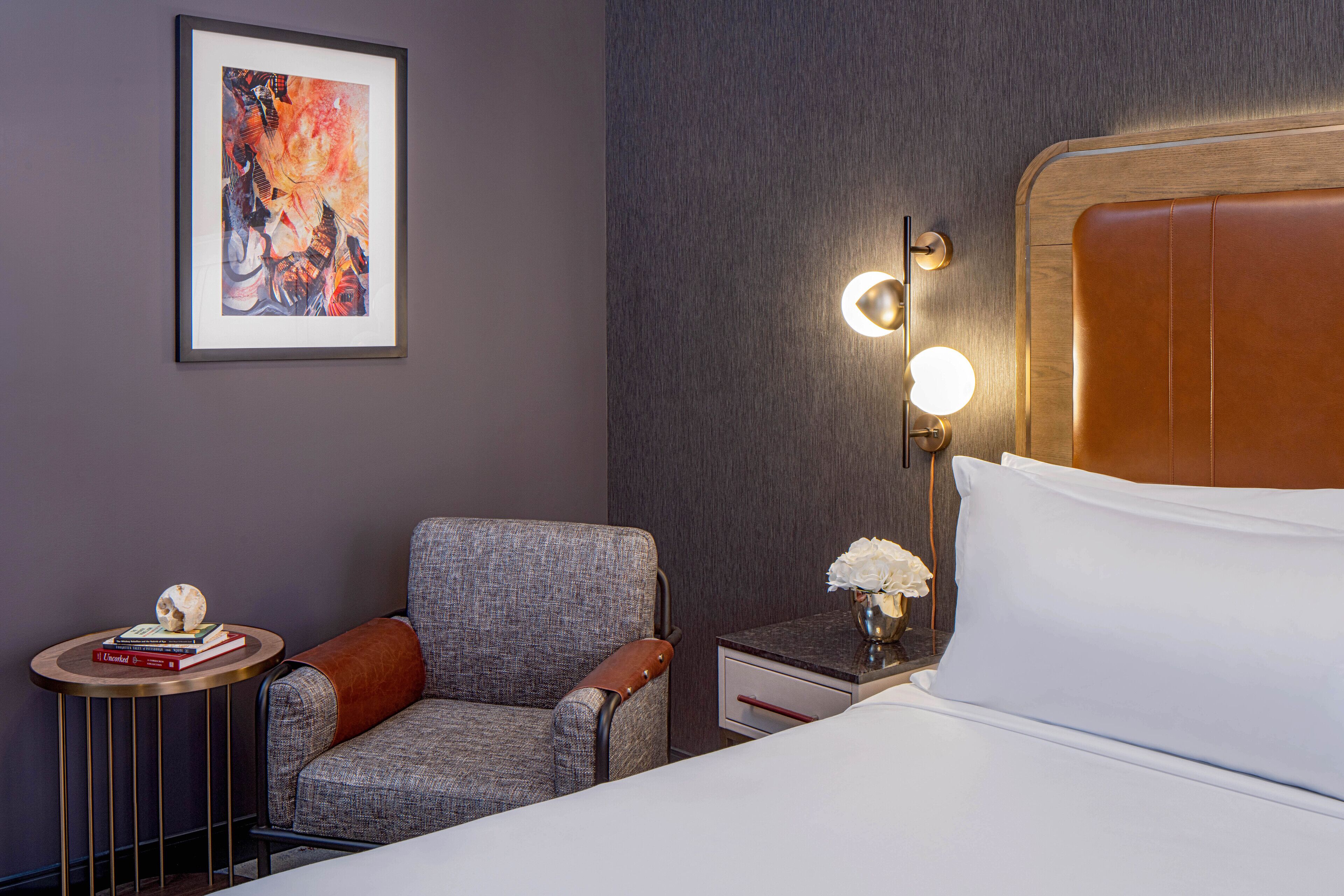 A hotel room with a bed, a chair, a nightstand, a framed picture, and a wall-mounted light fixture.