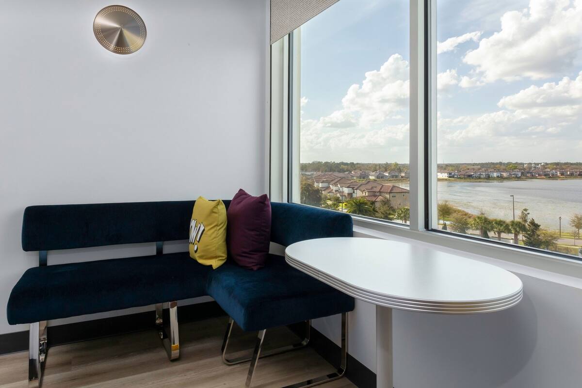 Una habitación con un banco azul oscuro, una mesita blanca pequeña y un ventanal con vistas a un río y edificios lejanos.