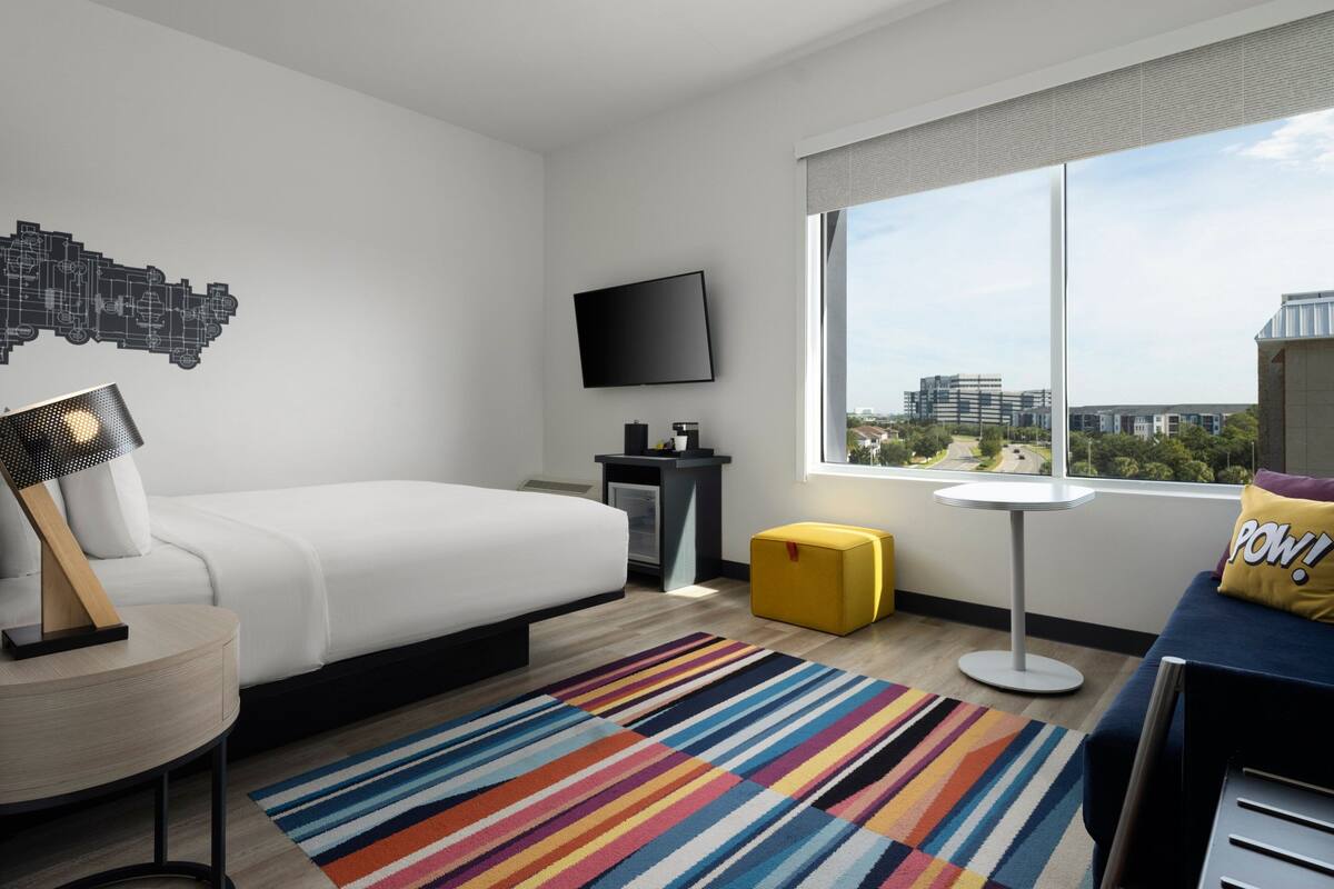 Una habitación de hotel moderna con una cama, un televisor, un escritorio pequeño, un tapete a rayas de colores y una ventana con vistas a la ciudad.