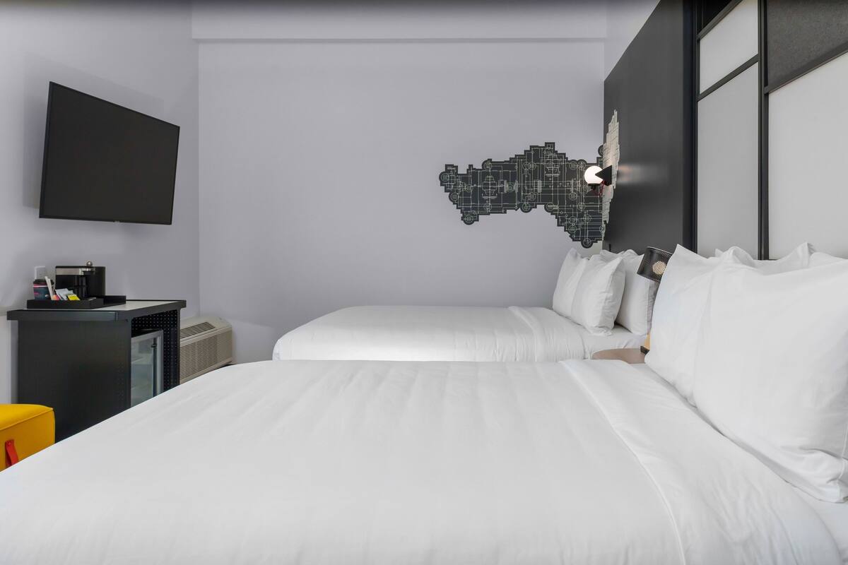 Habitación de hotel con dos camas, un televisor de pantalla plana, un escritorio con cafetera y una obra de arte en la pared.