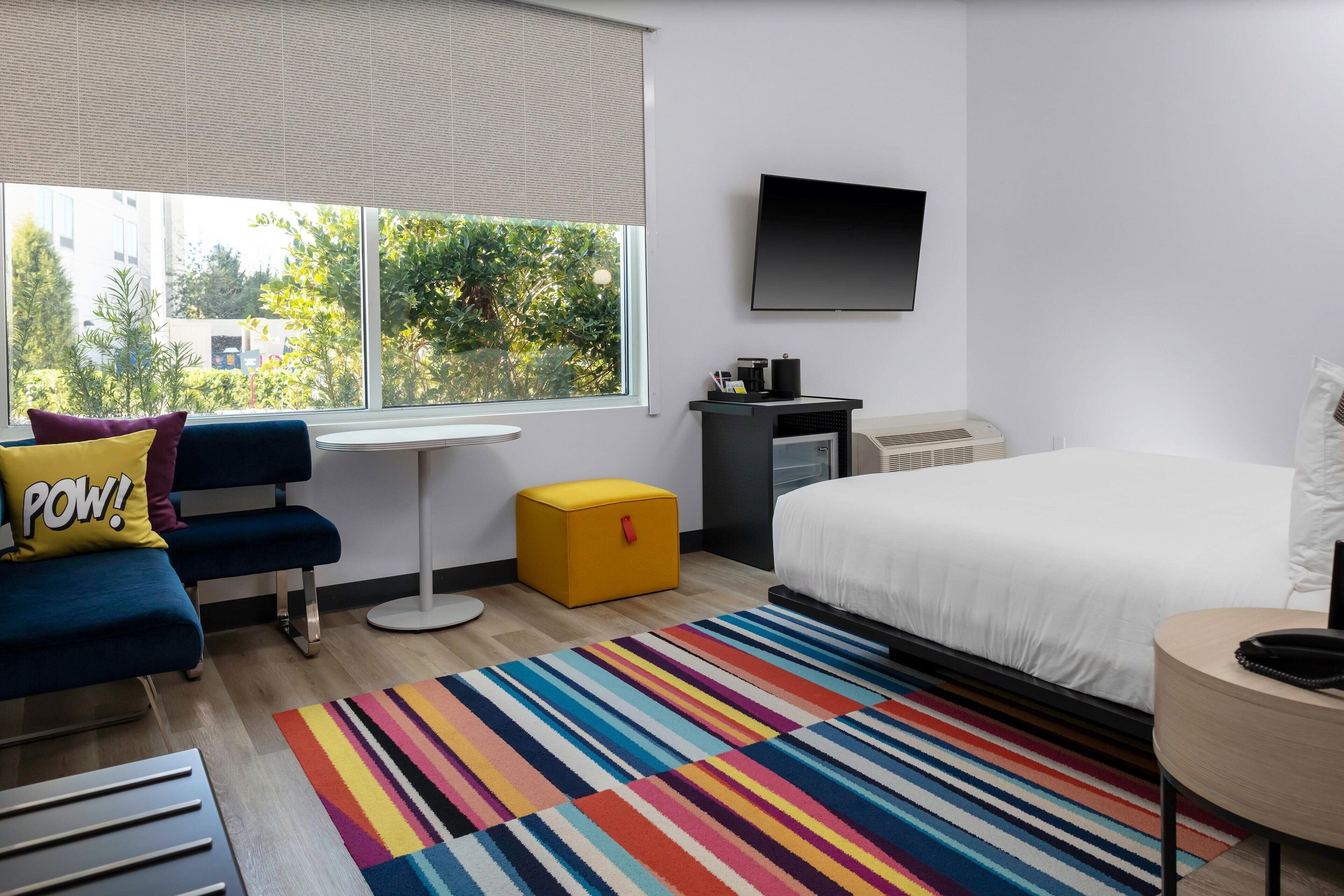 Una habitación de hotel moderna con una cama grande, un sofá azul con un cojín que dice 'POW!', un alfombra a rayas de colores, un pequeño taburete amarillo, un televisor de pantalla plana y una ventana con vistas a árboles y edificios.