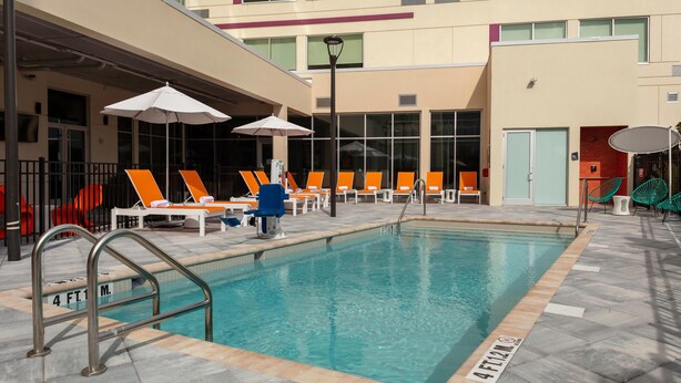 Une piscine avec des chaises longues et des parasols, une terrasse carrelée et un bâtiment doté de grandes fenêtres et d’une porte.