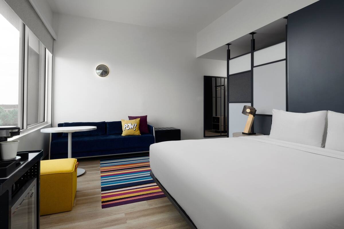 Una habitación de hotel moderna con una cama grande, un sofá, una mesita pequeña y una alfombra colorida a rayas.