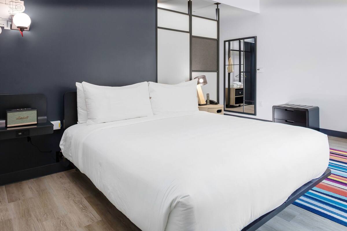 Una habitación de hotel moderna con una cama grande, una mesita de noche con lámpara, un espejo y una pequeña mesa auxiliar.