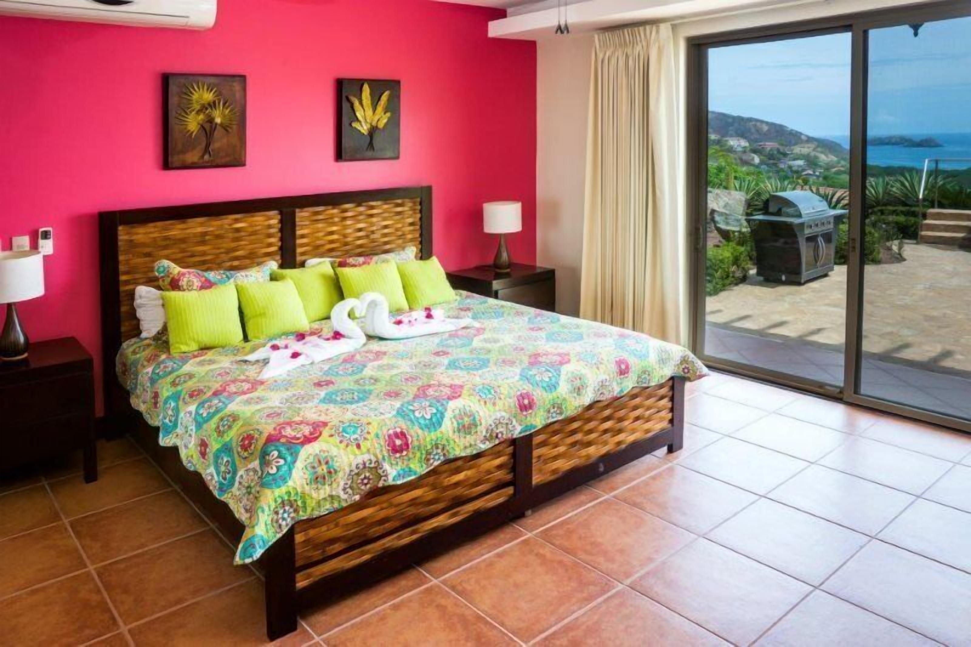 A bedroom with a large bed, a view of the outdoors, and a balcony with a grill.