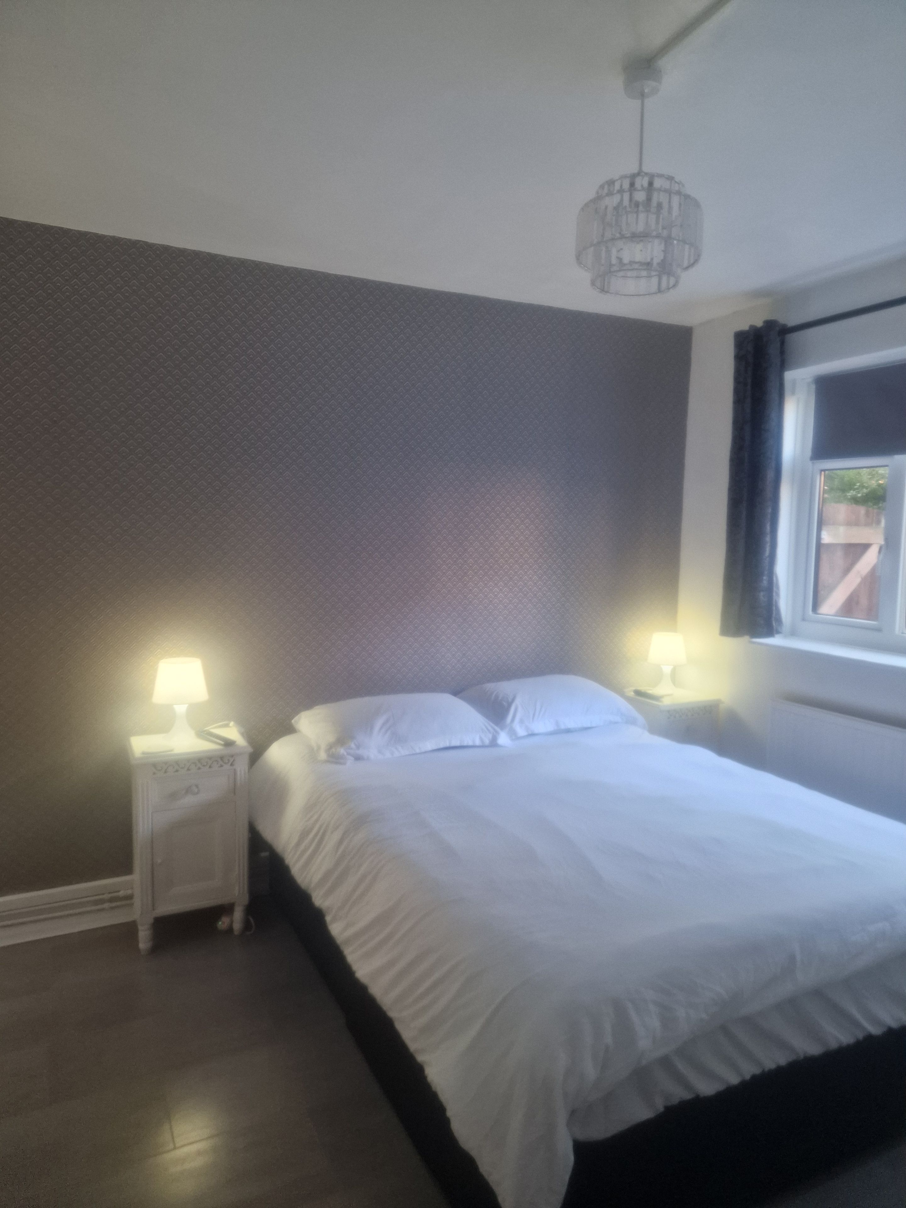 A bedroom with a bed, two bedside tables with lamps, a window with curtains, and a hanging light fixture.