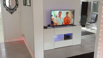 A modern living room with a flat-screen TV on a white entertainment unit, a black-framed mirror on the wall, and a black rug on the floor.