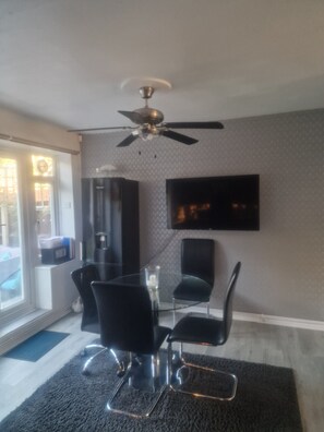 A modern dining area with a glass table, black chairs, a ceiling fan, and a flat-screen TV mounted on the wall.