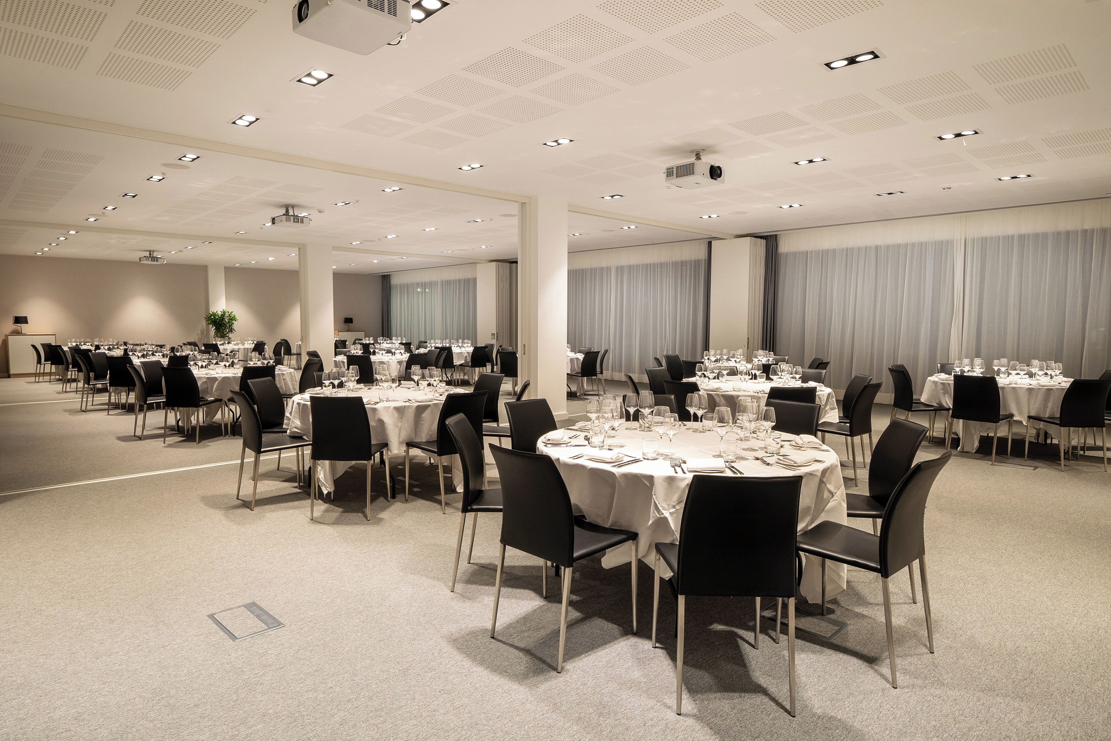 Un amplio salón de banquetos bien iluminado, con varias mesas redondas dispuestas para comer, sillas negras y manteles blancos.