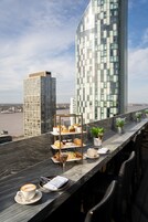 A terrace with a marble table set for a meal, including a coffee cup, pastries, and a wine glass, with a city skyline and river in the background.