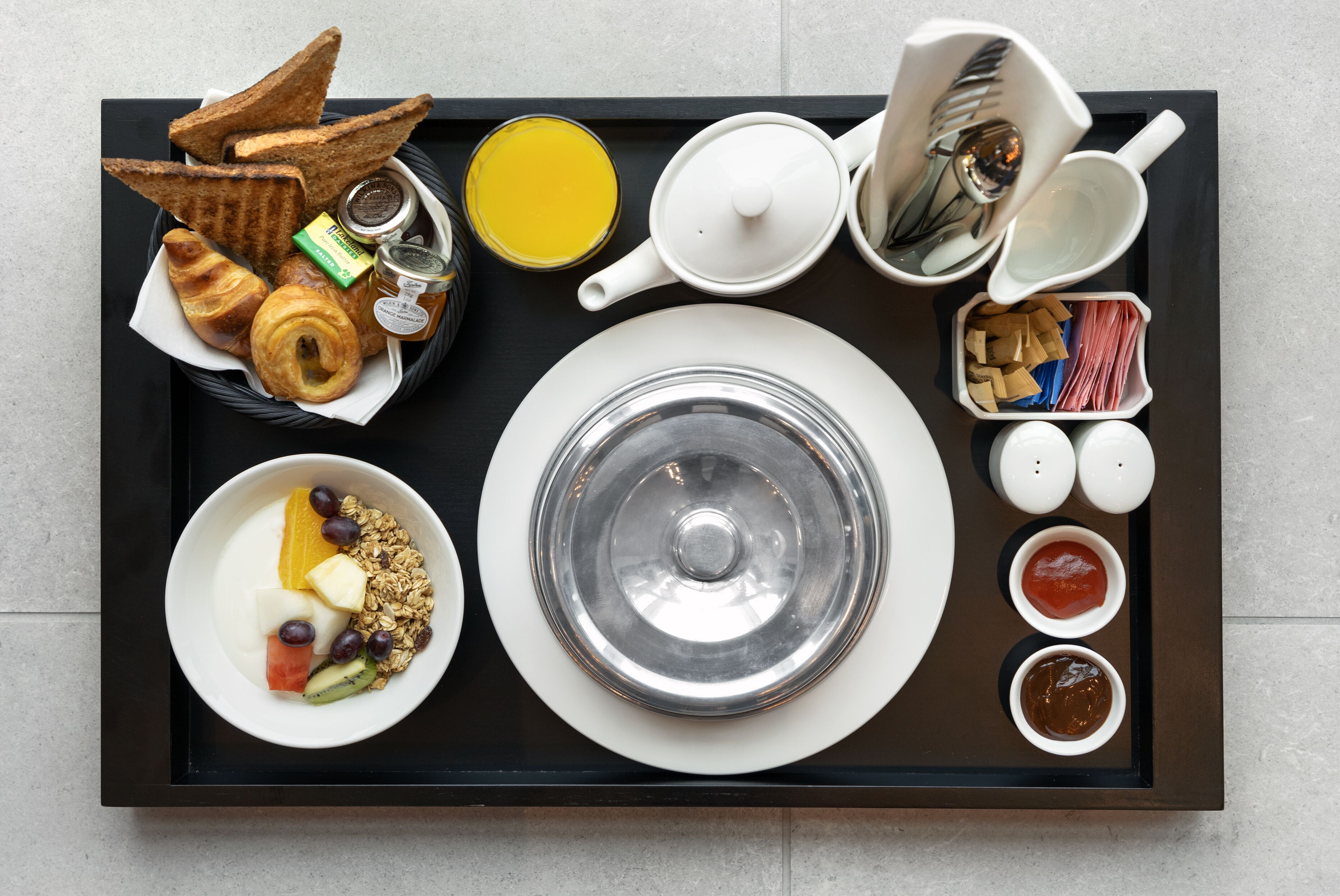Un plateau de petit-déjeuner avec des toasts, des croissants, du yaourt, du granola, des fruits et une théière.
