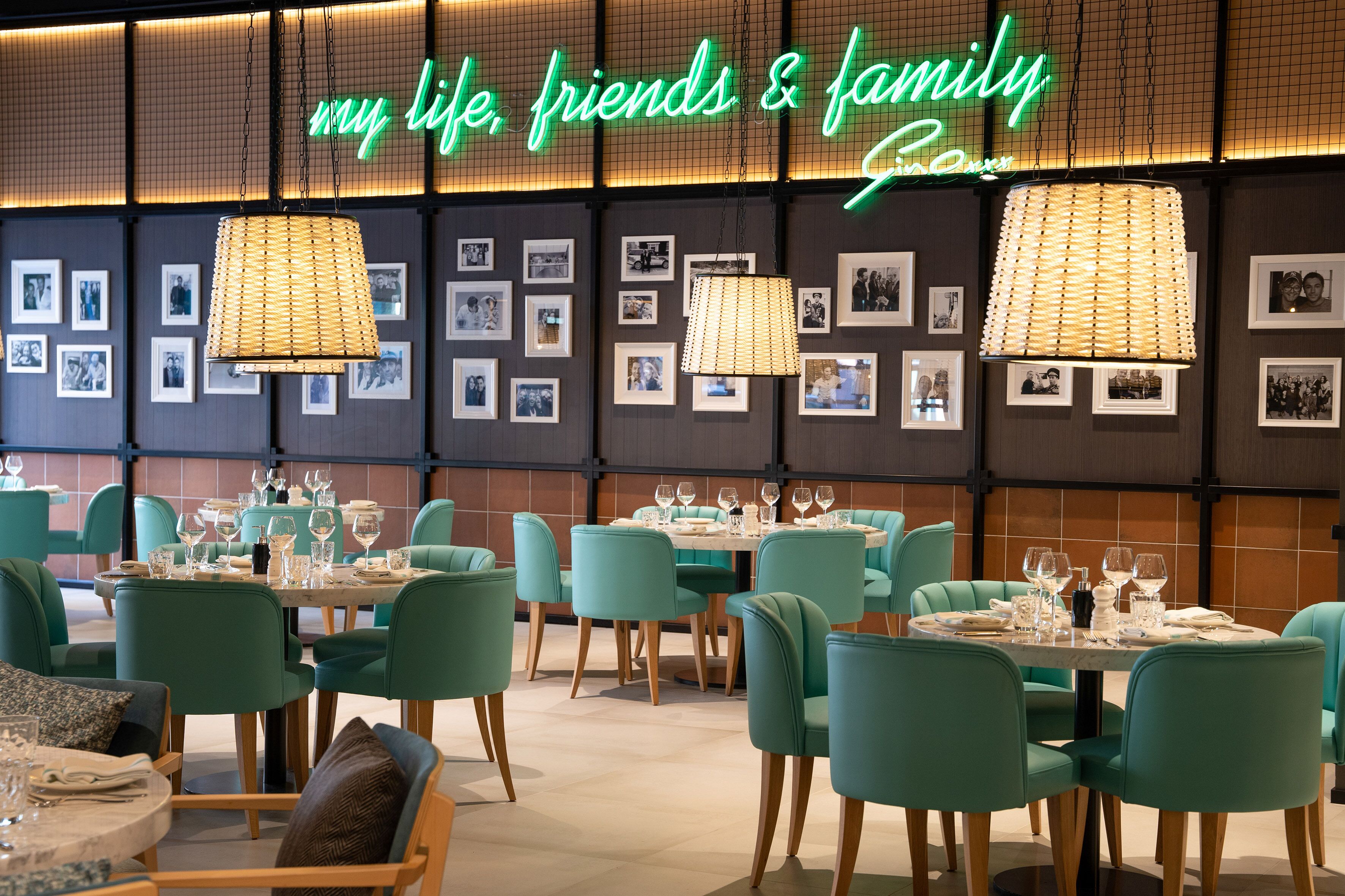 A restaurant with teal chairs and tables, framed photos on the wall, and a neon sign reading "my life, friends & family".