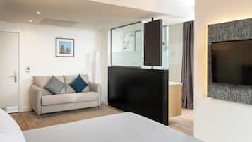 A modern hotel room with a large bed, a sofa, a TV, and a glass-enclosed shower area.
