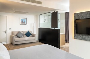 A modern hotel room with a large bed, a sofa, a TV, and a glass-enclosed shower area.