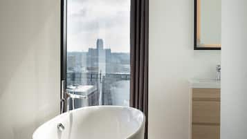 A modern bathroom with a freestanding bathtub, a large window with curtains, a wooden floor, and a minimalist sink area.