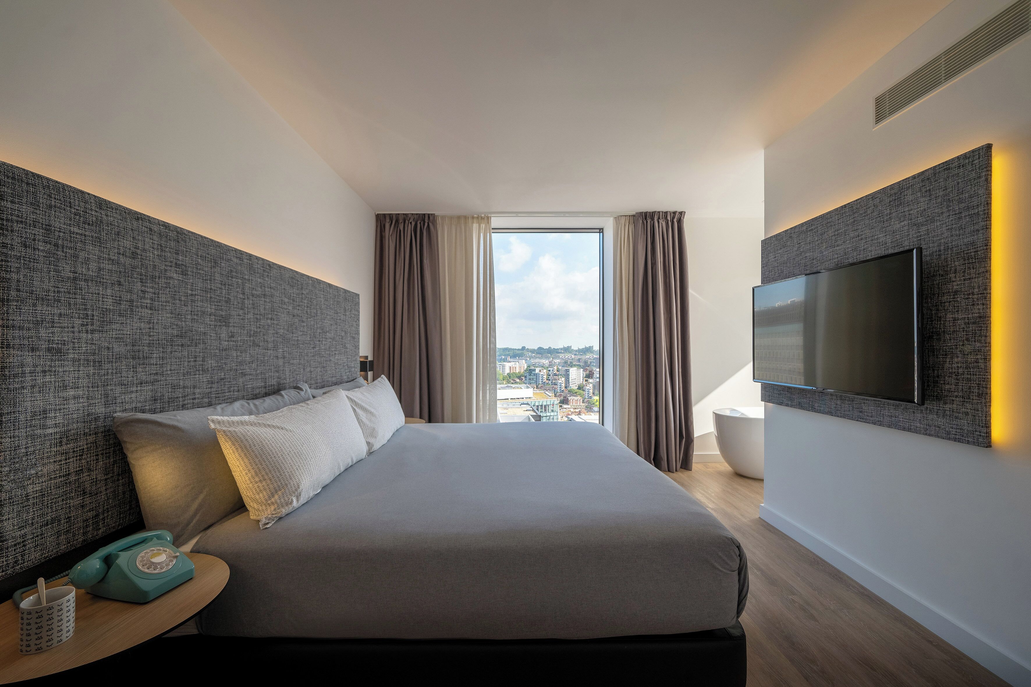 A modern hotel room with a large bed, a flat-screen TV mounted on the wall, a bedside table with a telephone, and a view of the city through the window.