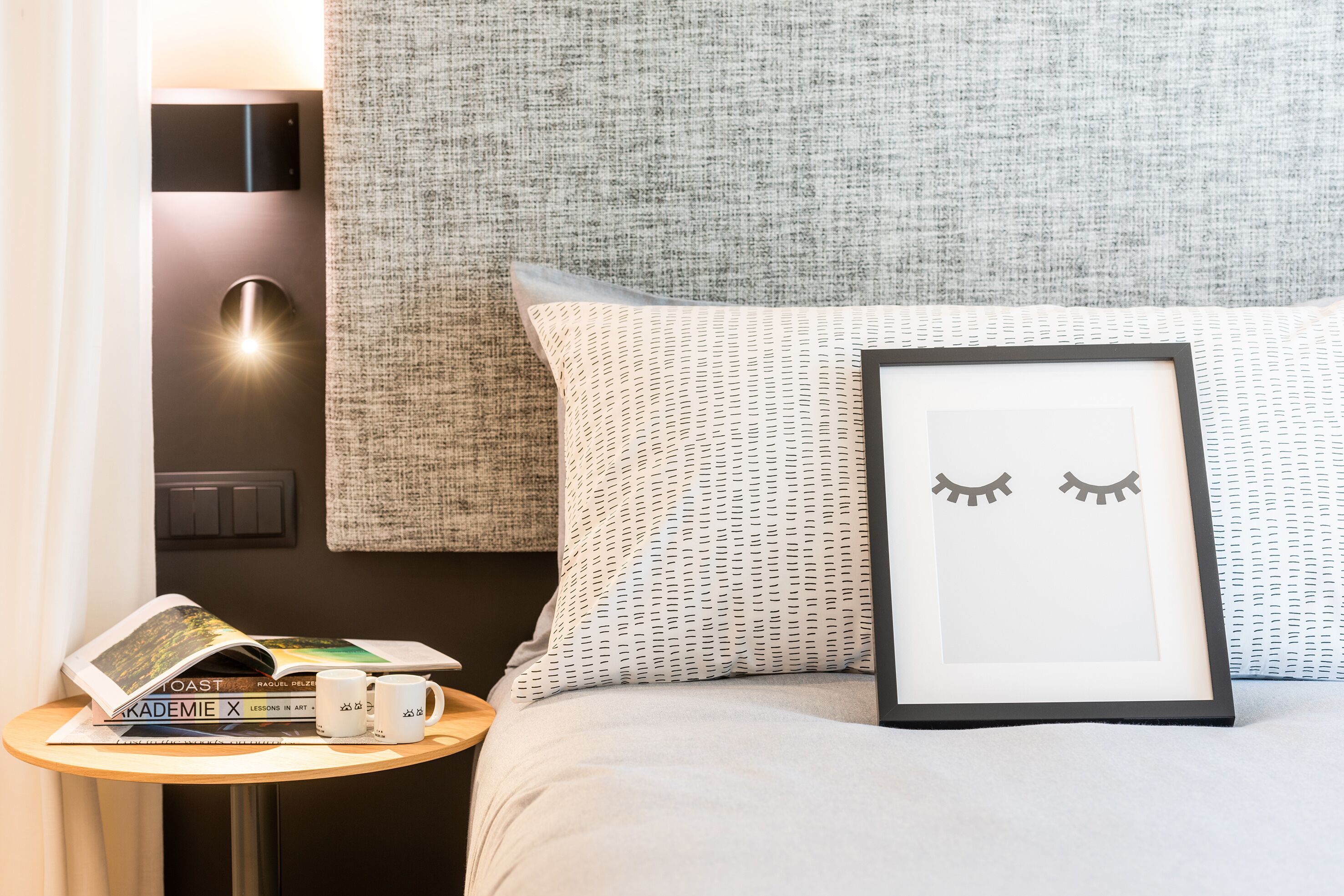 A neatly made bed with a framed picture of eyes, a small table with books and a mug, and a wall-mounted light.
