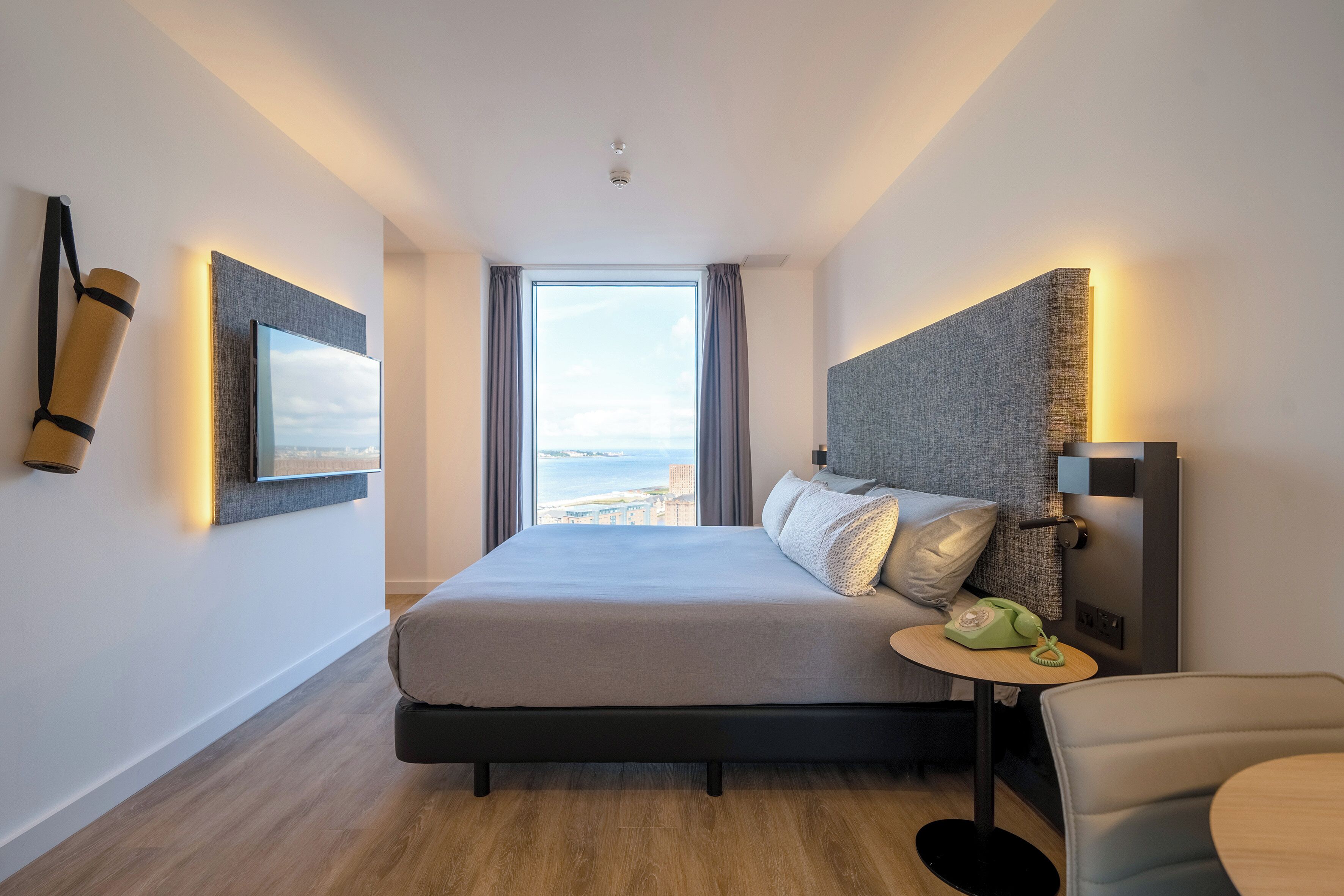 A modern hotel room with a large bed, a small table with a telephone, and a view of the sea.