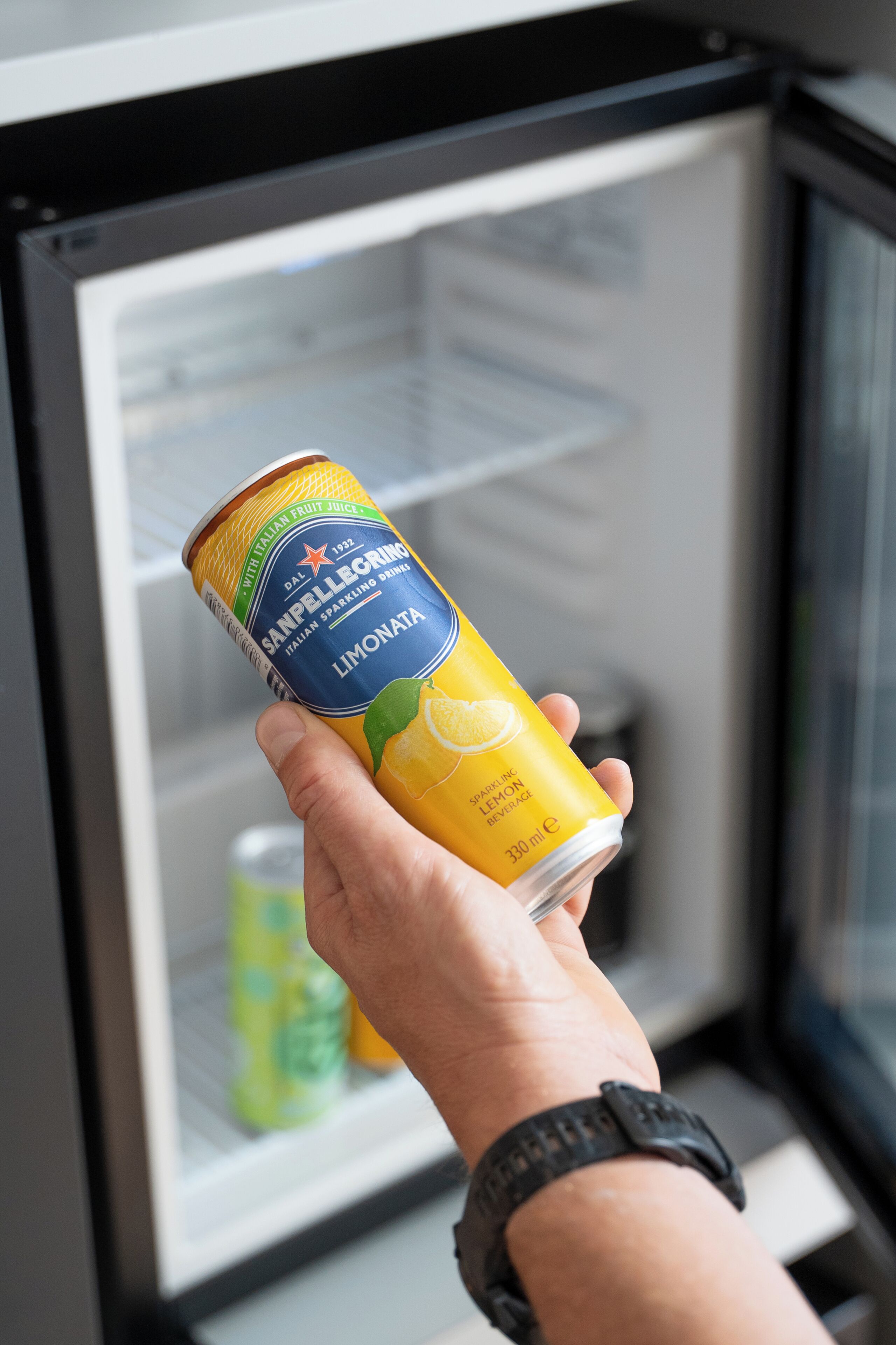 A hand holding a can of San Pellegrino Limonata Italian sparkling drink in front of a refrigerator.