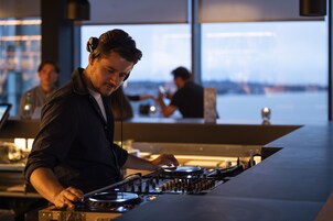A person operating DJ equipment in a modern setting with large windows.